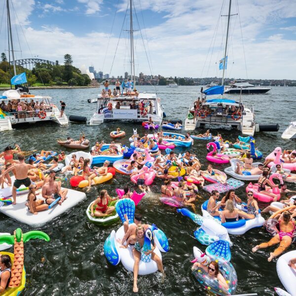 A large group of people enjoy a sunny day on a lake, surrounded by boats from The Yacht Social Club Event Boat Charters, with many floating on inflatable pool floaties shaped like unicorns, flamingos, and various other animals. The scene is lively and colorful, with numerous people in swimwear.