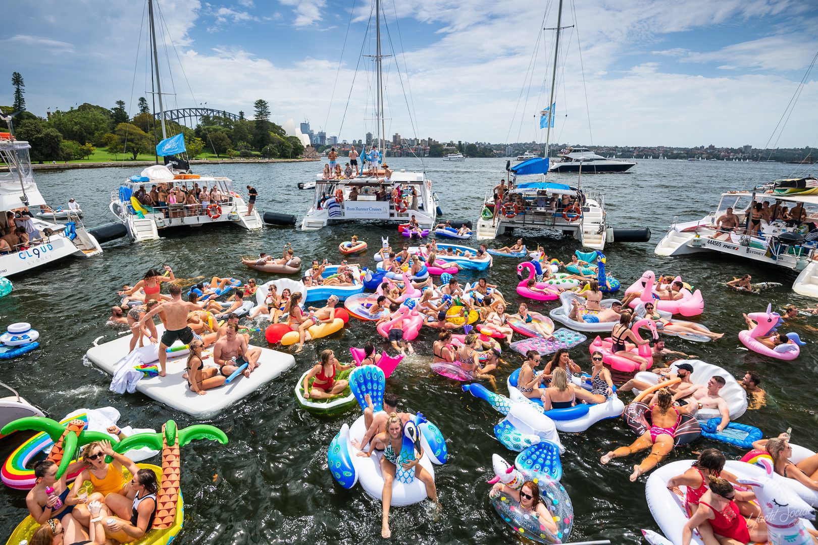 A festive scene on the water with numerous people on colorful inflatable rafts and pool floats. The Yacht Social Club Event Boat Charters are docked nearby, loaded with additional merrymakers. The sun is shining brightly, and the sky is partly cloudy. Everyone appears to be enjoying the fun atmosphere.