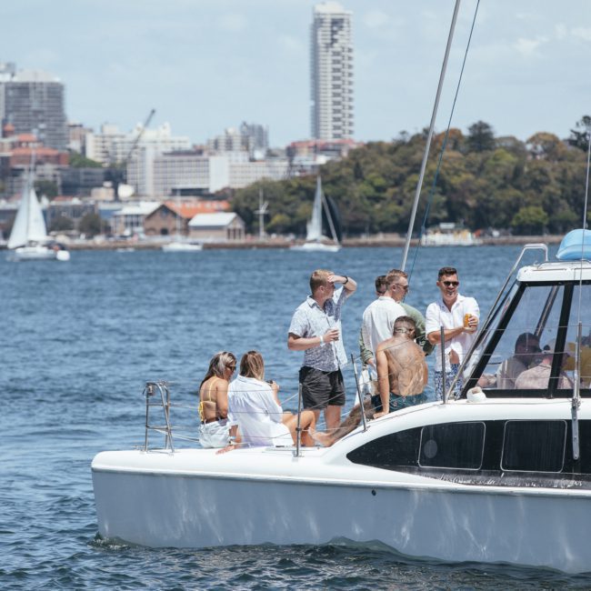 A group of people stands and sits on a catamaran in a harbor with city buildings and sailing boats in the background, enjoying a corporate boat event Sydney.
