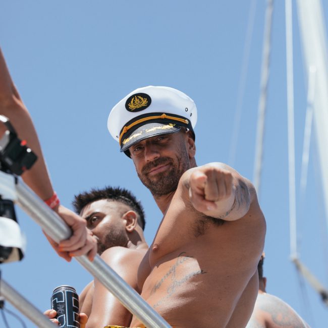 A shirtless man wearing a captain's hat stands on a boat, pointing at the camera during a lively Sydney boat party hire. Another man stands behind him, holding a can. The sky is clear and blue.