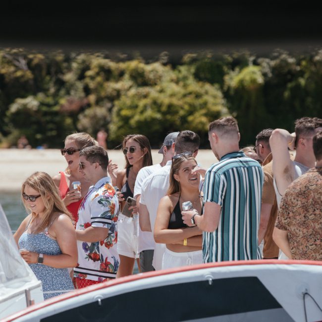 A group of people socializing on a beach near a luxury yacht, with some holding drinks. Dense foliage is visible in the background, perfect for those seeking Sydney boat party hire.