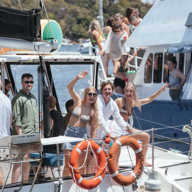 A group of people on a boat, smiling and waving. Another boat with people can be seen nearby. The background includes trees and part of a bridge, perfect for corporate boat events Sydney.