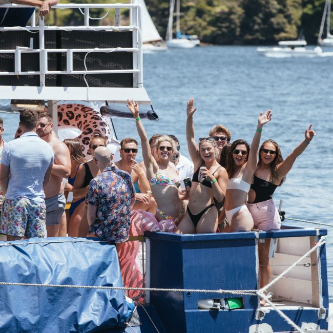 A group of people in swimsuits are standing and cheering on a luxury yacht anchored on a sunny day with sailboats and a forested shore in the background.