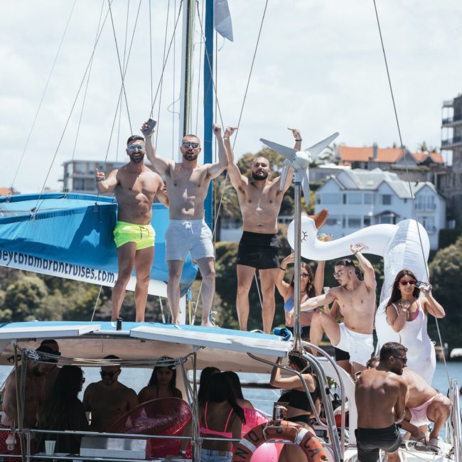 A group of people in swimwear is standing and sitting on a sailboat in a lively environment with buildings and trees in the background. Some people are waving and raising their arms, enjoying what appears to be a joyous Sydney boat party hire event.