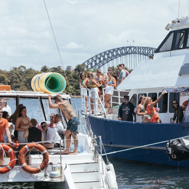 Two boats with groups of people on board are docked side-by-side on a sunny day, with the Sydney Harbour Bridge visible in the background—perfect for a corporate boat event or private yacht charter in Sydney Harbour.