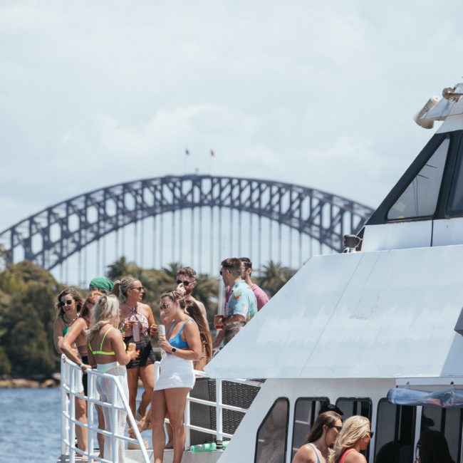 Individuals socializing on the deck of a boat with a large bridge visible in the background. The sunny day enhances their enjoyment of the view and each other's company, making it an ideal setting for luxury yacht hire Sydney.