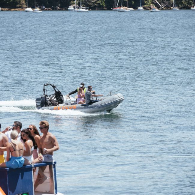 A small boat with filming equipment passes by people gathered on a dock by a body of water, with numerous sailboats in the distance. Ideal for corporate boat events Sydney or luxury yacht hire Sydney, this scene captures the essence of maritime charm.