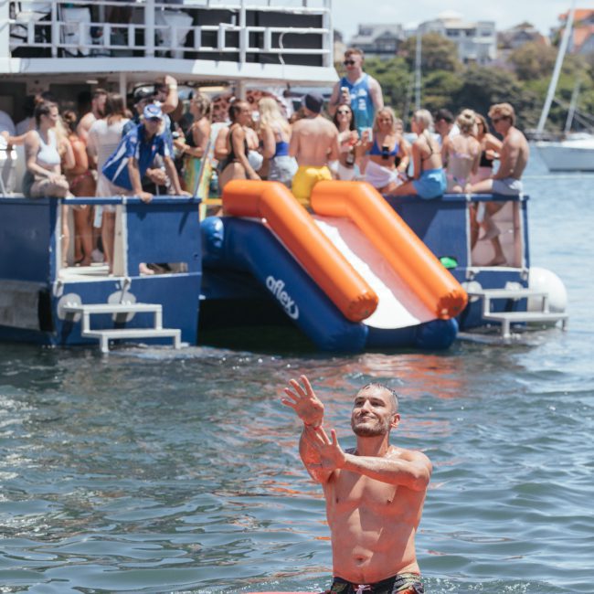 A shirtless man stands on a colorful inflatable in the water, reaching up. In the background, people are gathered on a boat with an inflatable slide extending into the water, highlighting the fun of a private yacht charter Sydney Harbour.
