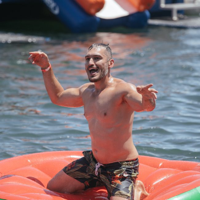 A man in swim trunks is kneeling on an inflatable raft in the water, raising his arms excitedly during a catamaran party Sydney.