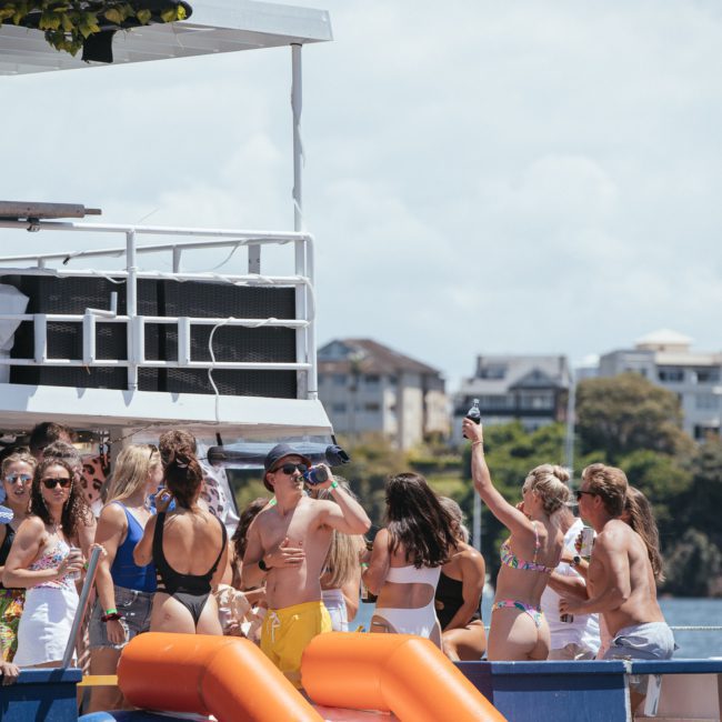 A group of people in swimwear are gathered on a luxury yacht, engaged in conversation and activities, with buildings and greenery visible in the background.
