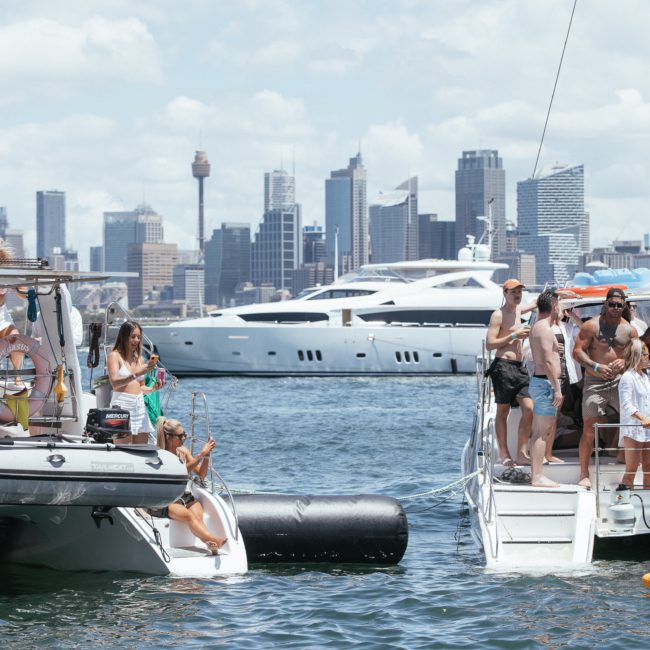 People enjoying a sunny day on yachts in a lively harbor with a city's skyline and large yacht visible in the background, embracing the vibrant scene of a catamaran party Sydney.