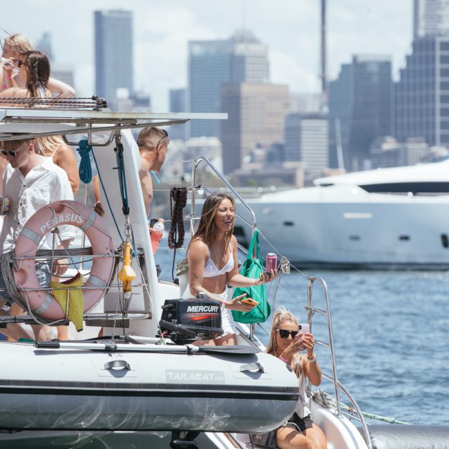 A group of people enjoying a sunny day on a private yacht charter in Sydney Harbour, with a city skyline in the background. Some are standing on the deck, talking and holding drinks, while others are seated on the boat's edge.