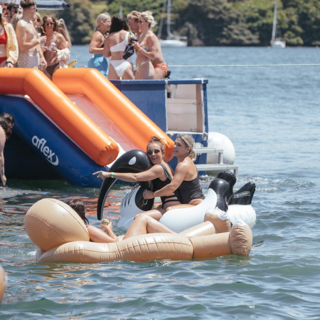 People enjoying a sunny day on a water slide and inflatable floats at a lake. Several are relaxing in the water, while others are on a dock near luxury yacht hire Sydney. Trees and boats add to the picturesque backdrop, enhancing the leisurely atmosphere.