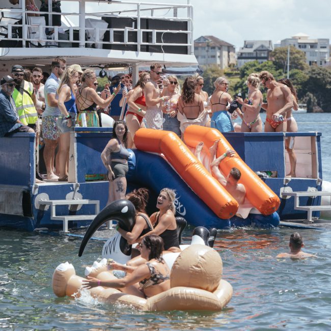 A group of people enjoy a sunny day on a catamaran party in Sydney with inflatable slides, while others play in the water with inflatable floaties. Buildings and trees are visible in the background.