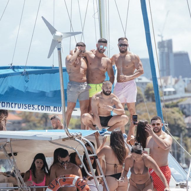 A group of people in swimwear stand and gather on a sailboat during a private yacht charter Sydney Harbour. Some are posing for the camera while others are engaged in conversation or relaxing on the boat, enjoying the vibrant atmosphere of their Sydney boat party hire.