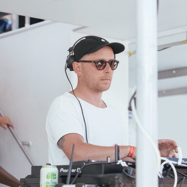 Man wearing sunglasses and a black cap operates DJ equipment on a table, with a white wall and staircase in the background. Perfect for your next private yacht charter on Sydney Harbour or corporate boat event.