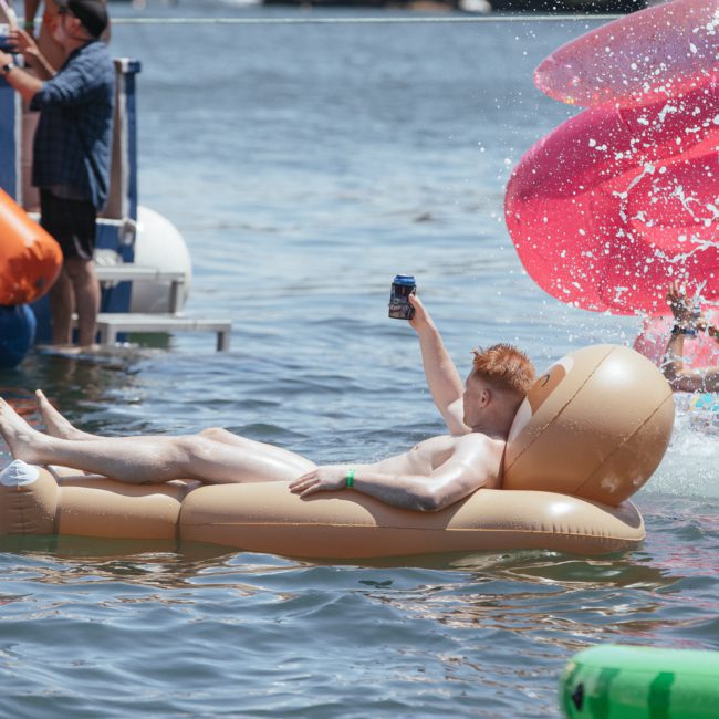 A person lounges on an inflatable chair in a body of water, holding a can up. In the background, other people enjoy inflatable structures during what appears to be a vibrant Sydney boat party hire event.