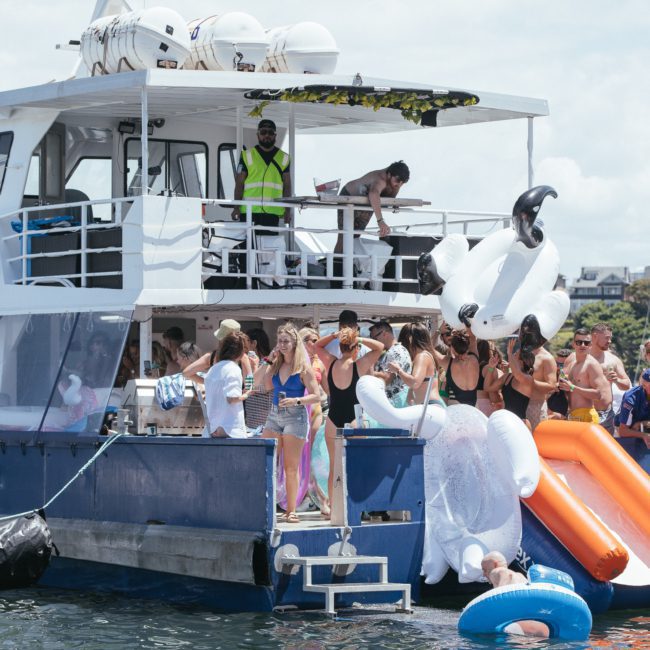 A group of people on the deck of a boat, some in swimwear, socializing and using inflatable toys, including a large swan float, enjoying a Sydney boat party hire.