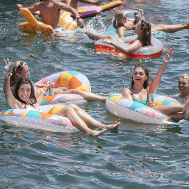 A group of people enjoying a sunny day, floating on colorful inflatables in a lake, waving and smiling at the camera as if they were on a Sydney boat party hire.