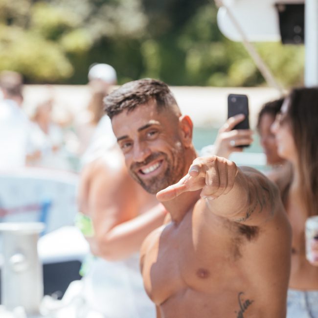 A shirtless man with tattoos points at the camera while smiling enthusiastically at an outdoor gathering. Several people can be seen in the background, enjoying the vibrant atmosphere of a Catamaran party in Sydney.