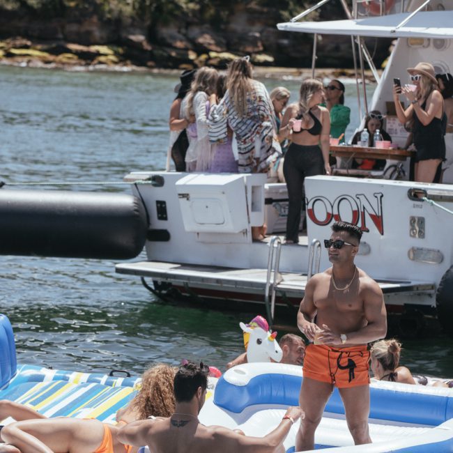A group of people are enjoying a sunny day on a boat and inflatable rafts in the water. Some are sitting and talking, while others socialize on a docked boat nearby, epitomizing the charm of private yacht charters in Sydney Harbour.