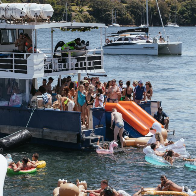 A group of people enjoy a sunny day on a dock with inflatable floats and slides, while others stay on the boat. Nearby, a catamaran party in Sydney sails on calm water with buildings visible in the background.