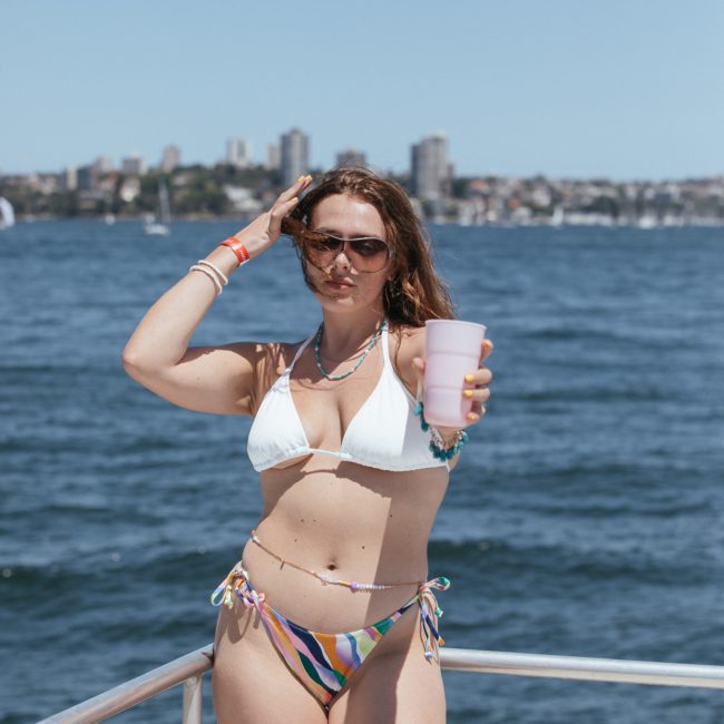 A woman in a white bikini top and patterned bottoms stands on a boat holding a pink cup. The ocean and Sydney cityscape are visible in the background, perfect for those considering a private yacht charter on Sydney Harbour.