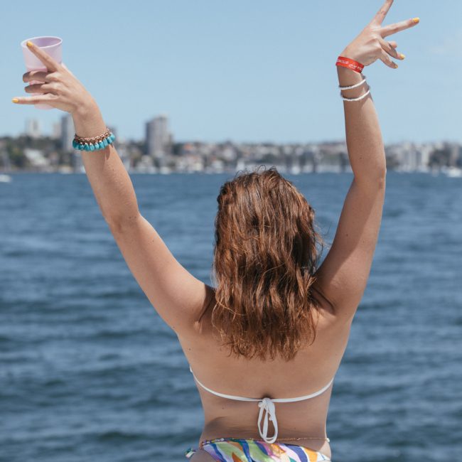 A person in a swimsuit stands on a luxury yacht hire Sydney, facing away, with arms raised, holding a cup, with a cityscape visible in the background.