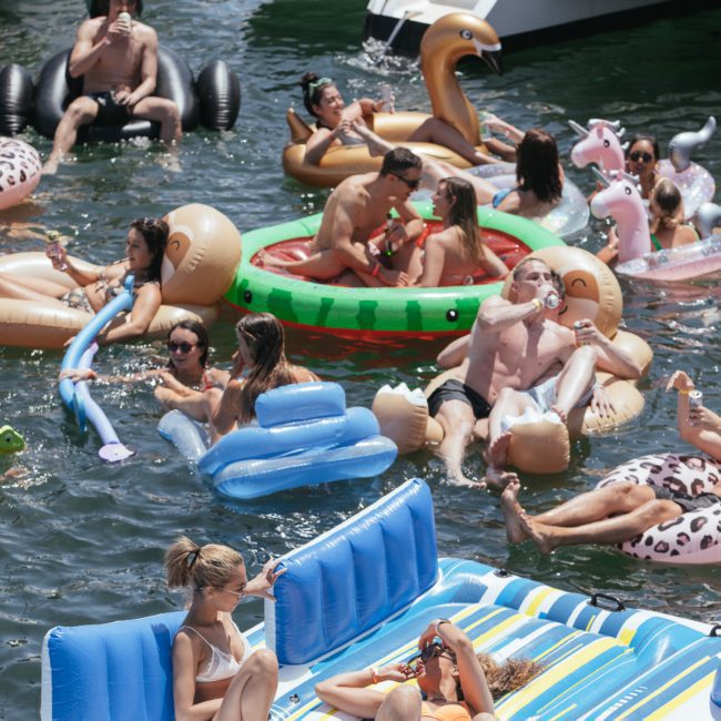 A group of people relax on inflatable rafts and floats in the water near yachts. They are enjoying a sunny day, chatting, and drinking. Perfect for a Sydney boat party hire — imagine the addition of a DJ boat hire Sydney to elevate the fun even more!