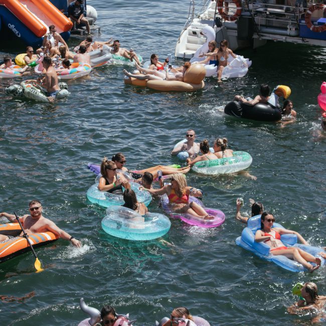 A group of people floating in a lake on inflatable pool toys near a luxury yacht. The scene includes various floaties, such as unicorns and donuts, with many enjoying the sunny weather. It looks like the perfect setting for a Sydney boat party hire.