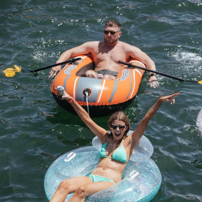 Two people floating on inflatables in a body of water. The man is in a gray and orange raft, while the woman in front is in a blue inner tube, smiling and raising her arms. It's reminiscent of the fun you could have at a Sydney boat party hire or catamaran party Sydney.