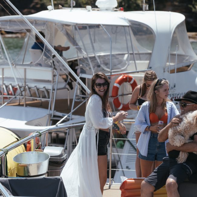 A group of people socialize on a private yacht charter in Sydney Harbour. One person holds a dog while others chat and enjoy drinks. The background shows another boat and greenery.