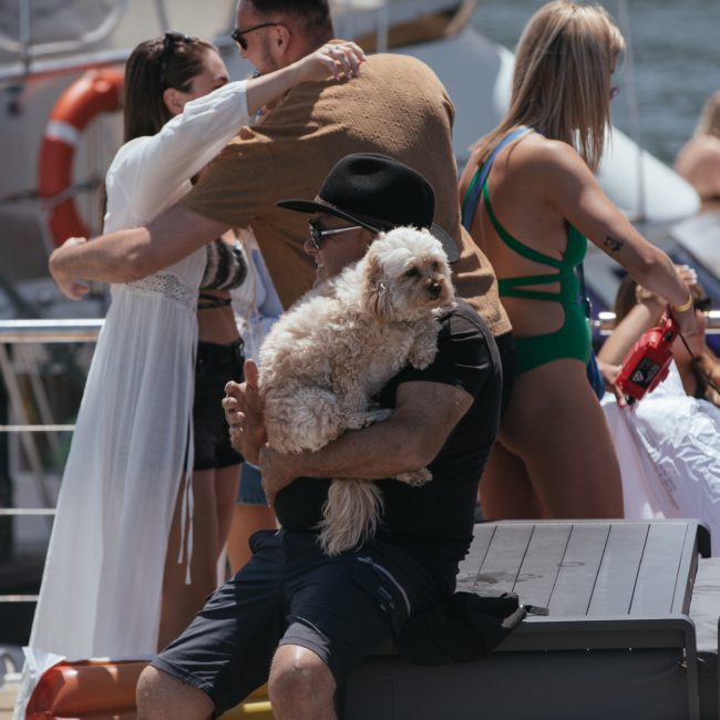 A man in a black hat and sunglasses sits on a boat deck holding a small white dog during a Sydney boat party hire. Several other people, some in swimwear, are socializing around him.