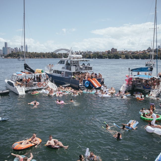A gathering of people on boats and inflatables in a body of water with a cityscape and bridge in the background under a partly cloudy sky, featuring luxury yacht hire Sydney.