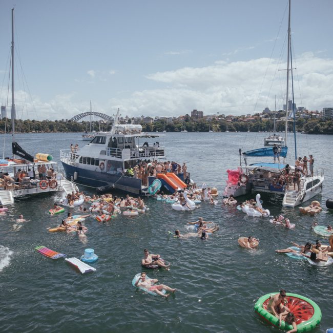 People enjoy a sunny day floating on inflatable items and swimming in the water near several boats docked together in Sydney harbor, making it feel like a Sydney boat party hire.