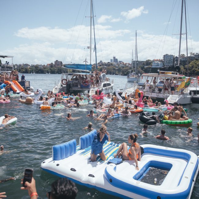 People on inflatable rafts and floats gather in the water near several anchored boats during a sunny Sydney boat party hire day.