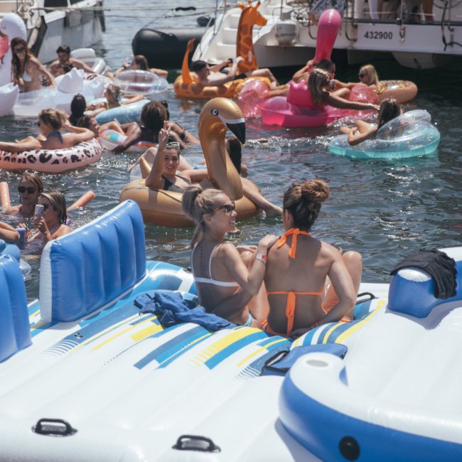 People are sitting on inflatable rafts and swimming while enjoying a sunny day on the water, surrounded by other inflatables and boats. It's the perfect setting for a catamaran party in Sydney, blending relaxation with vibrant energy.