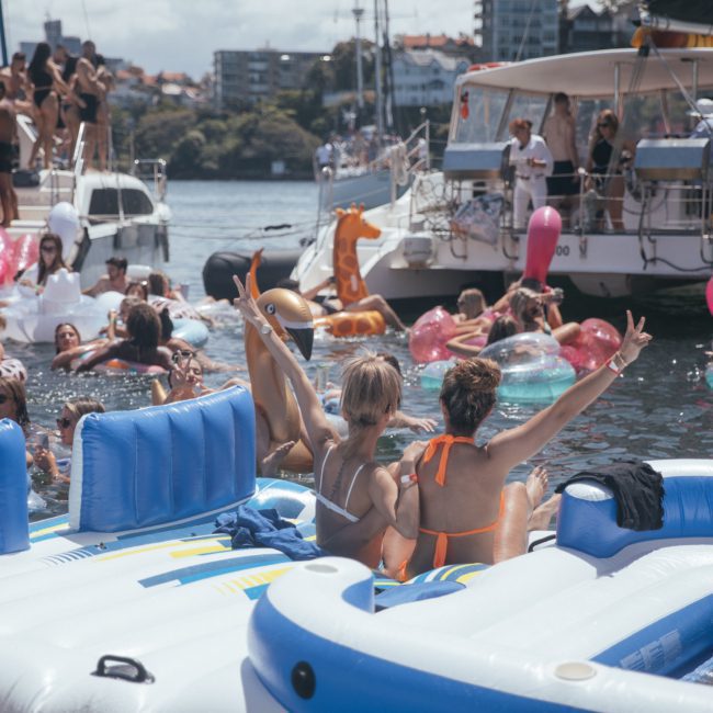 A crowd on various inflatable floats enjoys a sunny day in the water, with two women in the forefront making peace signs. Boats and a cityscape are visible in the background, hinting at the luxurious ambiance of a private yacht charter on Sydney Harbour.