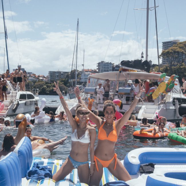 Two people are posing and making peace signs on inflatable floats in the water, surrounded by boats and other individuals on floats, with a backdrop of a sunny day and trees. Experience the fun with our Sydney boat party hire for your next event.
