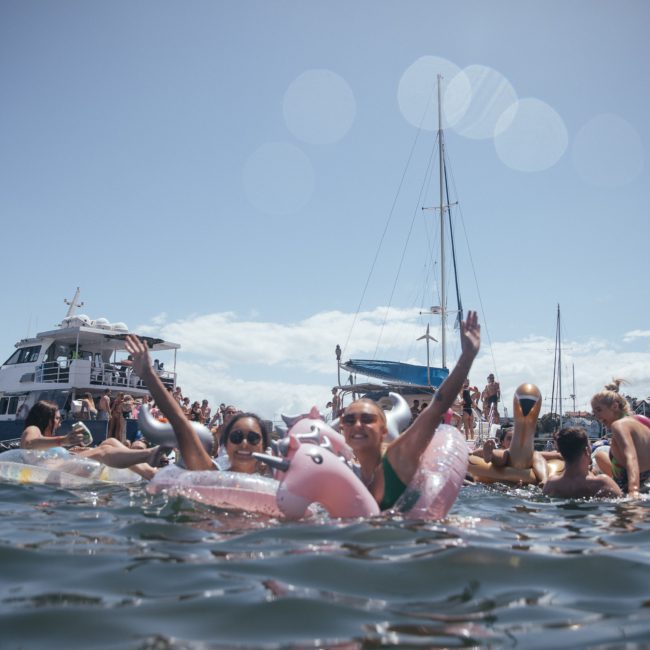 People are swimming in a body of water with inflatables, surrounded by boats on a sunny day, including a magnificent luxury yacht hire in Sydney.