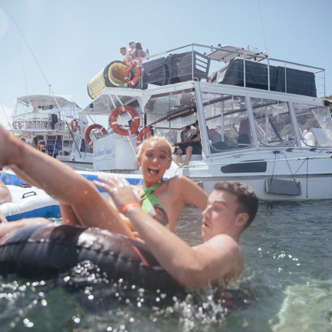 People enjoying a swim near boats on a sunny day, with one person laughing on a float while another plays in the water. A luxurious yacht hire in Sydney sets the perfect backdrop for this delightful scene.