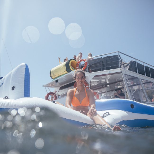 A person wearing an orange swimsuit sits on an inflatable raft in the water, with a houseboat and several people in the background on a sunny day. It's a perfect scene for someone enjoying a DJ boat hire Sydney experience.