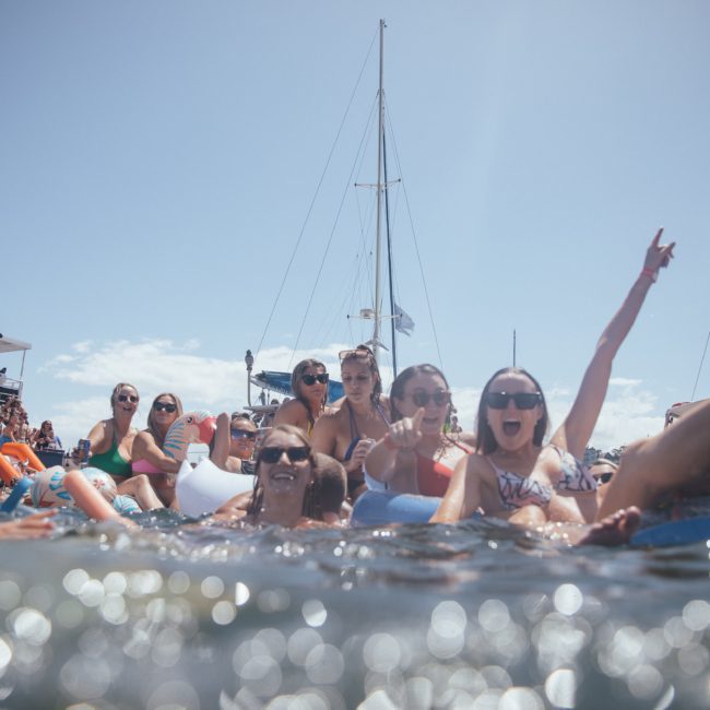 A group of people floats on inflatable tubes in the water near boats, under a clear sky. Some raise their arms and appear to be cheering and smiling, enjoying what looks like an exciting catamaran party Sydney thrill.
