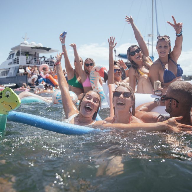 A group of people enjoying a sunny day in the water, surrounded by boats and inflatables, some raising their hands in excitement during a lively catamaran party Sydney.