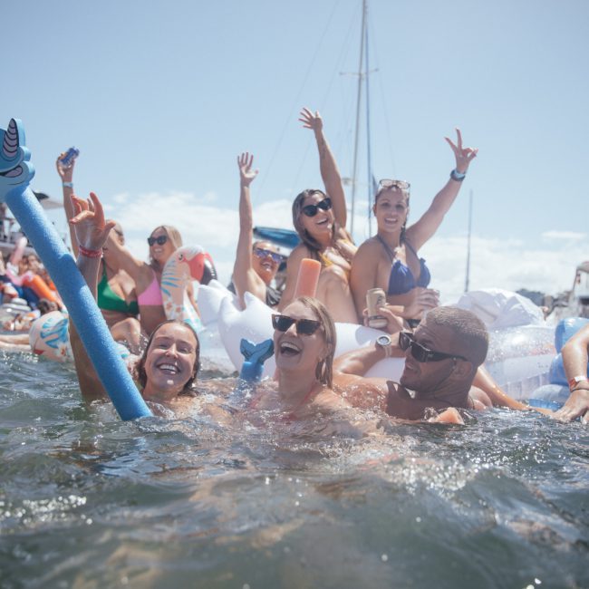 A group of people in swimsuits are enjoying a pool party with inflatables and boats in the background on a sunny day. Some are in the water smiling and raising their hands, making it feel like a Sydney boat party hire.