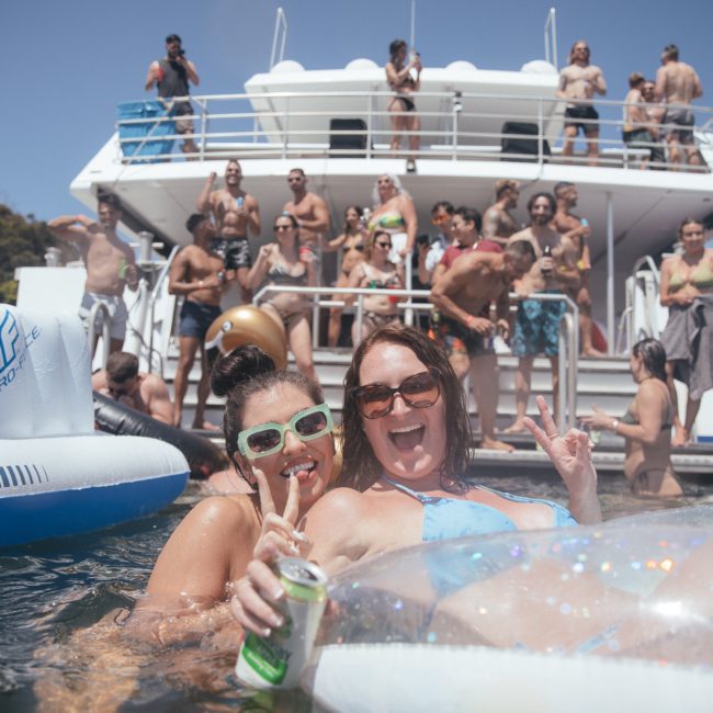 Two women enjoying a party on the water near a boat filled with people. One woman is holding a drink can and both are smiling and making peace signs while floating on an inflatable at a vibrant DJ boat hire Sydney event.