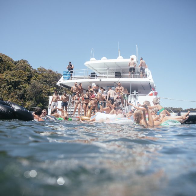 A group of people on a private yacht charter Sydney Harbour, enjoying a sunny day. Some are swimming while others are lounging on inflatable tubes. Trees are visible in the background.