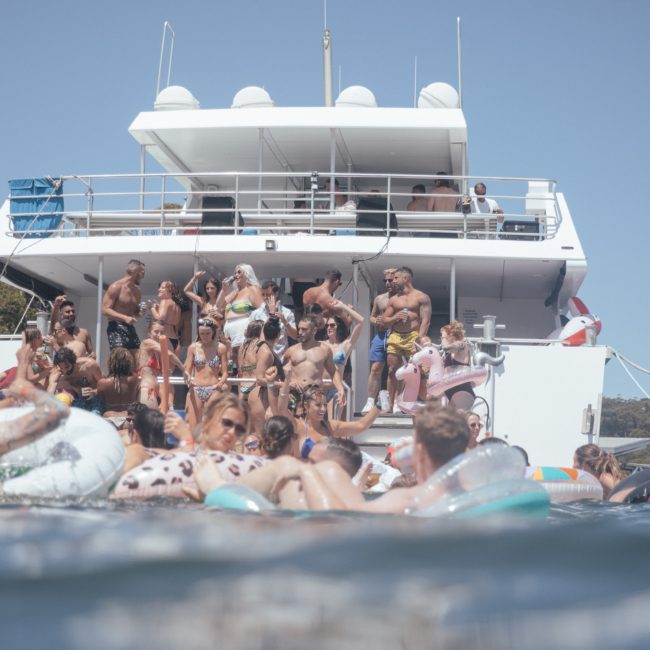 A group of people in swimsuits are enjoying a private yacht charter in Sydney Harbour. Some are on the upper deck, while others are swimming and floating on inflatables in the water. The yacht is anchored near a wooded shoreline, creating an idyllic setting for this luxury boat party.