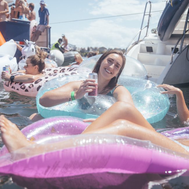 A group of people enjoys a sunny day on the water, lounging on inflatable rafts and boats, with one woman in the foreground smiling and holding a drink. Consider a luxury yacht hire in Sydney for an even more unforgettable experience.