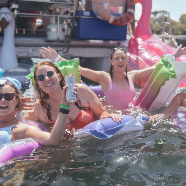 Four women in swimsuits are enjoying drinks while floating on inflatable rafts in a body of water with a private yacht charter on Sydney Harbour in the background.
